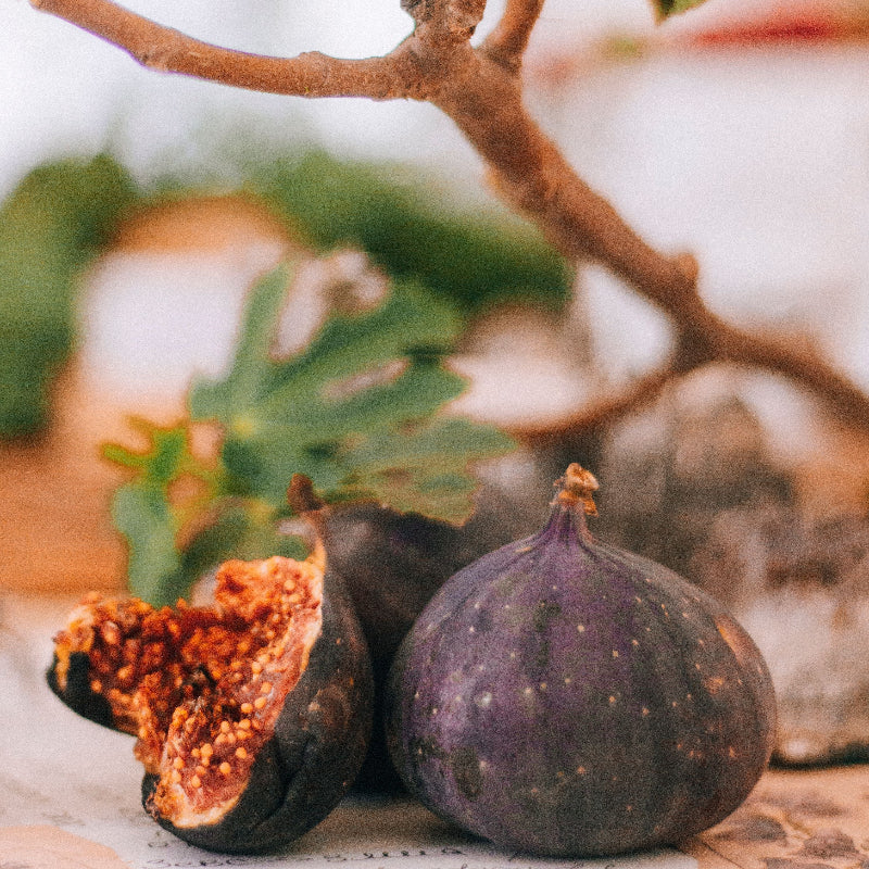Fresh figs, including one whole dark purple fig and two halved figs showing orange-red seeds, arranged on a stone surface with a fig branch and green leaves in the background.